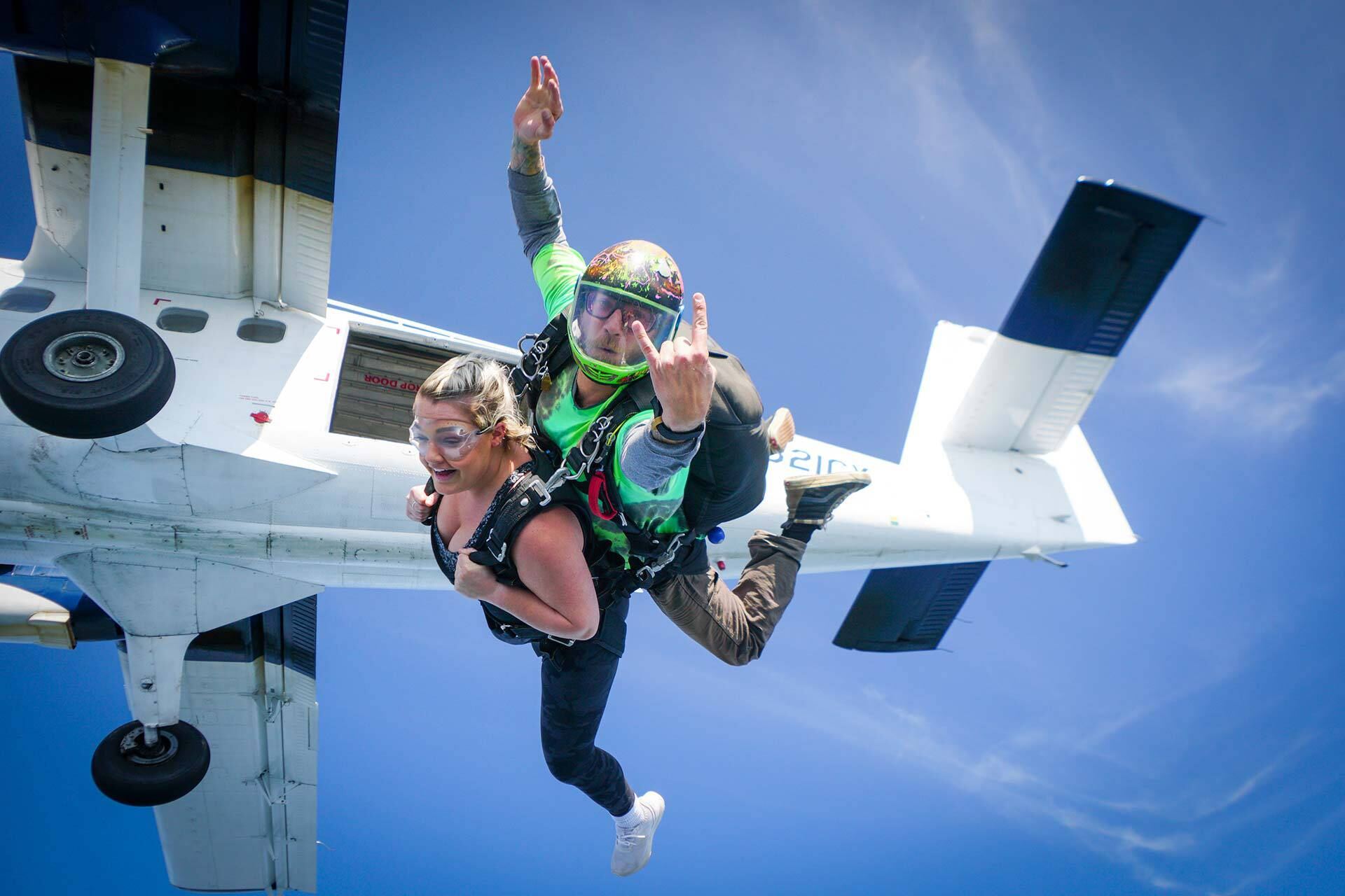 Una coppia in tandem esce da un aereo da paracadutismo, la passeggera sorride con gli occhiali e le braccia aperte
